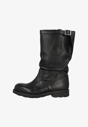 Musta nahkaiset polven korkeudelle ulottuvat saappaat, joissa on taitettu varsi, metallinen solkivivahde ja teksturoitu pinta. Nelikulmainen kärki ja paksu, tukeva pohja.