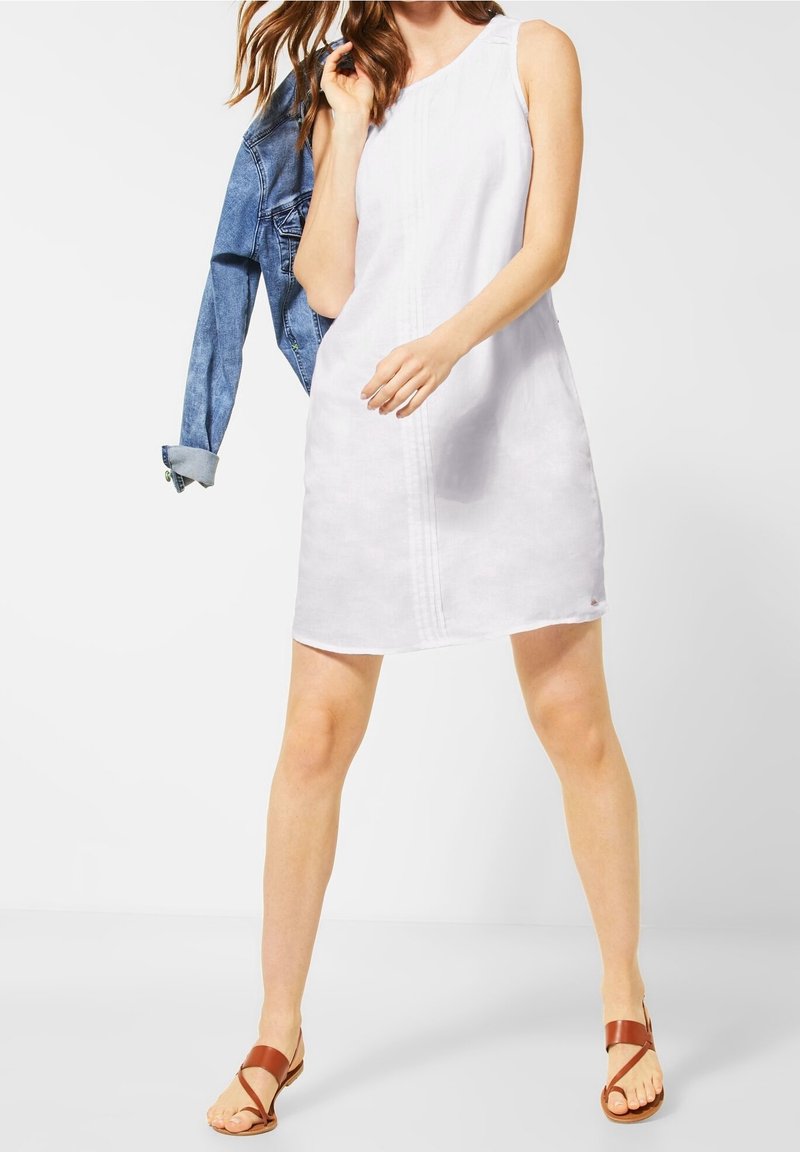 Femme portant une robe blanche sans manches et des sandales marron tient une veste en jean bleue sur l'épaule sur un fond uni.