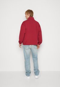 Giacca pullover bordeaux con colletto alto, vestibilità rilassata, spalle scese, abbinata a jeans azzurri chiari e sneakers bianche. Design semplice.