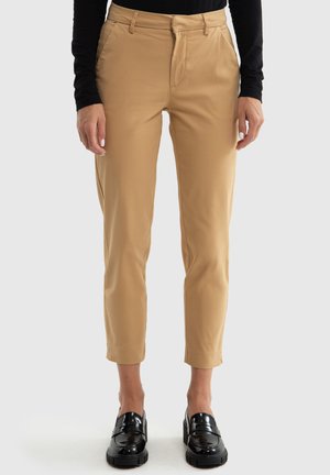 Beige maßgeschneiderte Hose mit schlanker Passform, flacher Front, seitlichen Taschen und schmal zulaufendem Bein. Getragen mit schwarzen glänzenden Slippern.
