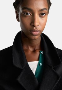 Cappotto di lana nera con colletto ampio, indossato sopra un cardigan verde con bottoni blu, su una camicia bianca.