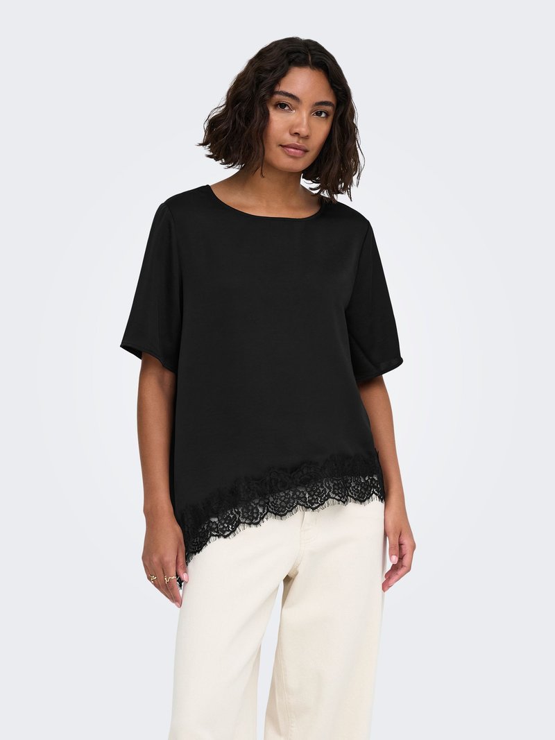 Femme aux cheveux bouclés mi-longs, portant un haut noir à manches courtes avec bordure en dentelle et un pantalon écru, debout devant un fond uni.