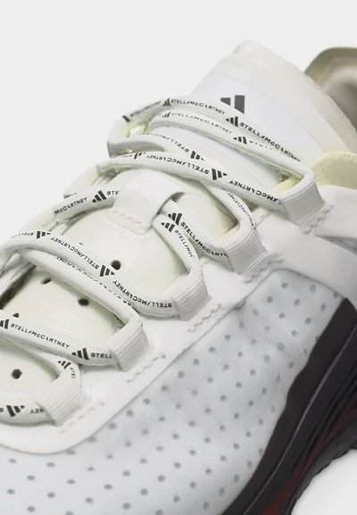 Bela superga z perforiranimi stranicami, belimi vezalkami z napisom "Stella McCartney" in logotipom Adidas na jezičku.