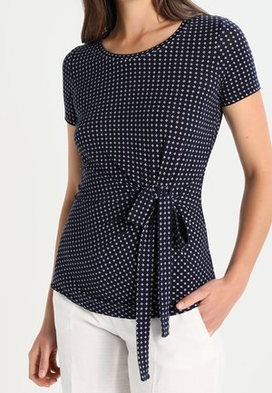 Mujer con una blusa de manga corta azul marino con pequeños patrones de cruces blancas y un cinturón atado a la cintura, combinada con pantalones blancos.