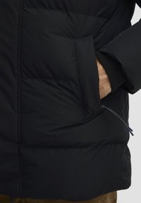 Sort puffergæk med quiltet design, der har en sidelomme, elastiske manchetter og en lynlåsgreb med en blå accent.