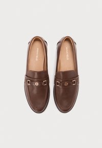 Mocasines de cuero marrón con una textura suave, que presentan herrajes decorativos y un sutil acento de logo en el empeine. Diseñados con una punta redonda.