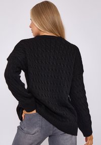 Schwarzer Strickpullover mit strukturiertem Muster, weitem Ärmel und gerippten Bündchen. Die Rückseite hat einen asymmetrischen Saum, kombiniert mit grauen Jeans.