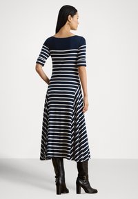 Γυναίκα που στέκεται με το χέρι στη μέση, φοράει μεσάτο φόρεμα με navy  και άσπρες ρίγες, μανίκια μέχρι τον αγκώνα και μαύρα μπότες με τακούνι, κοιτάζοντας πλευρικά.