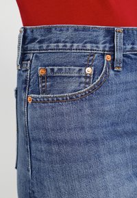Närbild av en blå denimjeansficka med orange sömnad och kopparnitar mot en röd tygbakgrund.