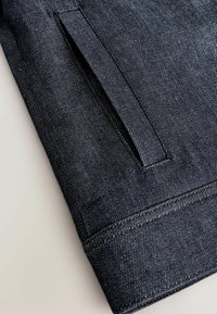Tume sinine denim kangas, millel on rafineeritud tekstuur, küljetasku ja kontrastne valge õmblus, mis jookseb õmbluste ääres.