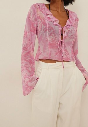 Różowa przezroczysta bluzka w róże, z falbankowym dekoltem i wiązaniem z przodu. Długie rękawy z rozszerzanymi mankietami, w zestawie z białymi, wysokim stanem spodniami.