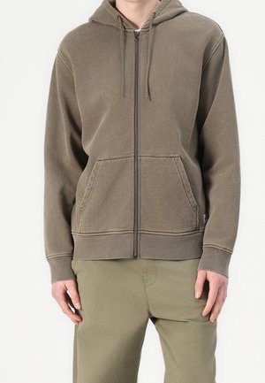 Mann trägt einen braunen Kapuzenpullover mit Reißverschluss und Fronttaschen sowie passende olivgrüne Hose, steht mit entspannten Händen an den Seiten.