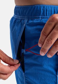 Blue shorts with an elastic waistband, featuring a zippered pocket with a red drawstring inside. Material appears lightweight and textured.