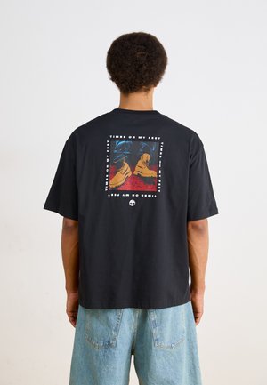 Black cotton t-shirt with a rectangular graphic design featuring boots against a colored background. Short sleeves and a relaxed fit.