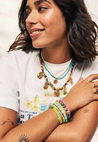 Goldfarbene und farbige Perlenketten übereinander getragen auf einem T-Shirt, mit bunten Perlenarmbändern an einem Arm, der ein kleines Blumentattoo zeigt.