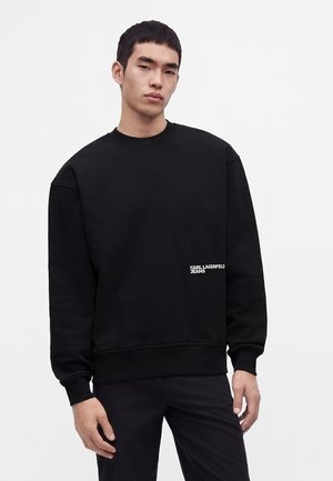Młody mężczyzna w czarnej bluzie z długim rękawem z logo Karl Lagerfeld Jeans przy talii, stojący na jednolitym tle.