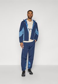 Chaqueta cortavientos en azul marino y azul con capucha, combinada con pantalones a juego. Presenta un diseño texturizado y puños elásticos, llevada sobre una camiseta de rayas.