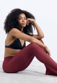 Schwarze Spitze-Bralette und burgunderfarbene Leggings, getragen von einem Modell, das auf einer hellgrauen Oberfläche sitzt und glatte Texturen sowie ein figurbetontes Design präsentiert.