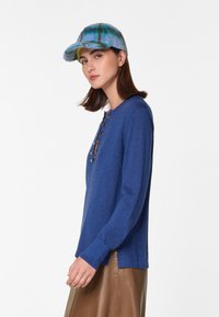 Blauwe gebreide top met een ronde halslijn en decoratieve knopen. Gecombineerd met een bruine leren rok en een kleurrijke pet.