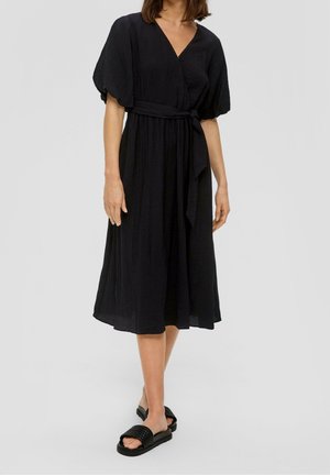 Femme portant une robe mi-longue noire à manches bouffantes et ceinture à la taille, accompagnée de sandales noires à enfiler sur un fond uni.