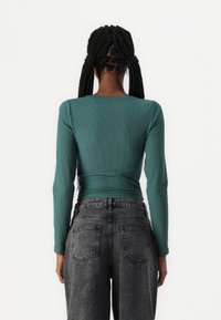 Langermet ribbestrikket topp i teal, med rund hals og en tettsittende silhuett, kombinert med høy midje mørkegrå jeans.