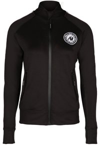 Chaqueta negra con cremallera y cuello alto, que cuenta con dos bolsillos laterales con cremallera y un parche de logo circular en el lado izquierdo del pecho. Textura de tela suave.