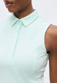 Gros plan d'une personne portant un polo sans manches bleu clair avec un petit logo blanc sur la poitrine gauche.