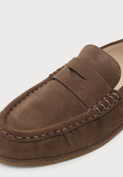 Mocassin marron en daim avec détail de coutures visibles et une décoration fendue sur le dessus, présenté sur un fond clair uni.