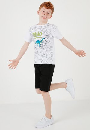 Ragazzo dai capelli rossi che indossa una maglietta bianca con stampa di dinosauri, pantaloncini neri e scarpe da ginnastica bianche, in equilibrio su una gamba con le braccia distese.