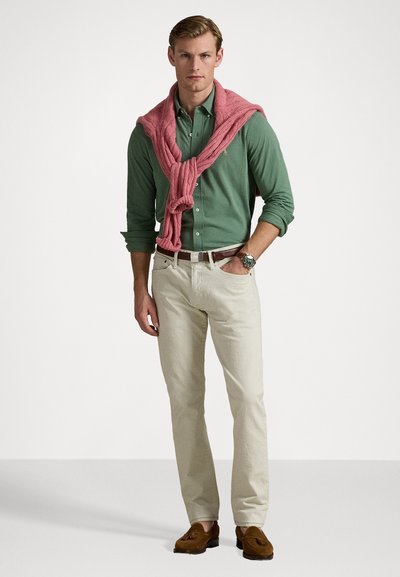 Manlig modell i en grön skjorta med knappar, krämfärgade chinos, bruna loafers och en rosa stickad tröja över axlarna.