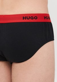 Schwarze knappe Unterhose mit einem kontrastierenden roten elastischen Bund, der den Text "HUGO" zeigt. Glatte Textur und körpernahe Passform.