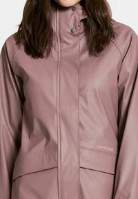 Rosa vattentät jacka med hög krage, tryckknappar och sidofickor. Slät yta med synliga sömmar. Logotyp på sidan.