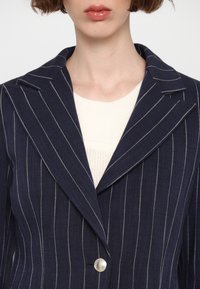 Femme portant un blazer bleu marine à fines rayures avec de larges revers sur un top en maille blanc, visible du cou jusqu'au milieu du torse.