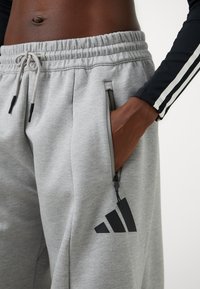Pantaloni tuta grigi con cintura elastica e cordino, caratterizzati da tasche con zip nere e da un logo Adidas nero sulla gamba sinistra.