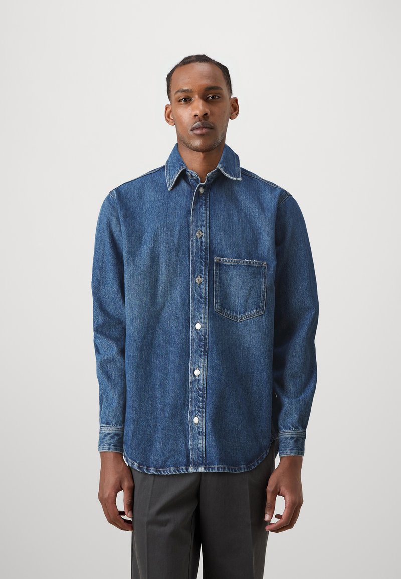 Camicia di jeans di colore blu scuro, maniche lunghe, colletto a punta, taschino frontale e chiusura con bottoni. Presenta un orlo consumato e una texture robusta.