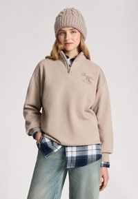 Beżowa polarowa bluza z półzamkiem, z logo. Noszona na niebieską koszulę w kratę i połączona z jasnymi dżinsami. Gruba dziana czapka.