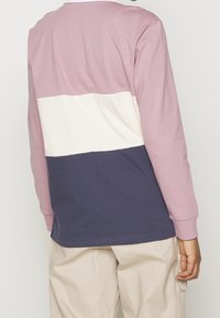 Person, der ein langärmliges, mehrfarbiges Hemd mit mauve, weiß und navy Abschnitten sowie einer hellbeigen Hose trägt, von hinten gezeigt.