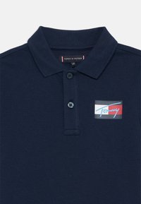 Mörkblå pikétröja i texturerad bomull, med krage, två knappar och ett färgglatt logopatch på vänster sida.