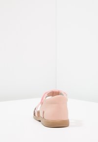 Różowe sandały dziecięce z rzepem i brązową podeszwą, pokazane od tyłu na białej powierzchni i neutralnym tle.