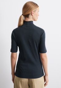 Juodas trumpas rankovės polo marškinėliai su imitaciniu apykakle, gobaliniu tekstūru ir prigludusia siluetu, derinami su béžinėmis kelnėmis.