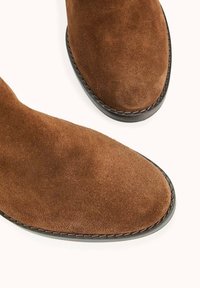 Bottines en daim marron avec une texture lisse, un bout arrondi et une semelle en caoutchouc noire contrastante. Aucune quincaillerie ni motif visible.