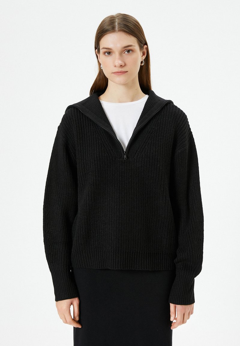 Schwarzer gestrickter Pullover mit einer Kapuze mit Reißverschluss, ausgestattet mit einem strukturierten Muster und gerippten Bündchen. Getragen über einem weißen Oberteil und einem schwarzen Rock.