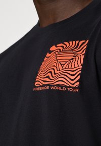 Zwarte katoenen T-shirt met een gestileerde oranje grafiek van golven en bergen, met de tekst "FREERIDE WORLD TOUR" eronder.