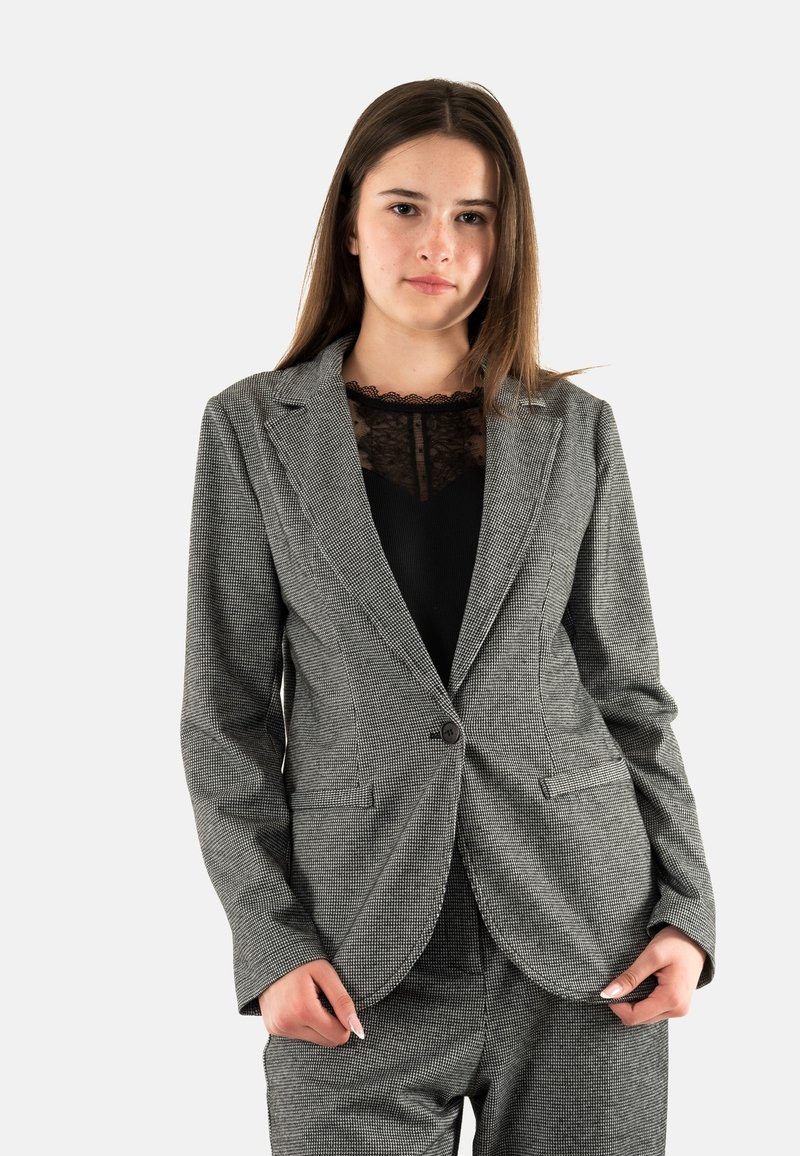 Blazer de cuadros gris con hombros estructurados, cierre de un botón y dos bolsillos frontales, llevado sobre una blusa de encaje negra.