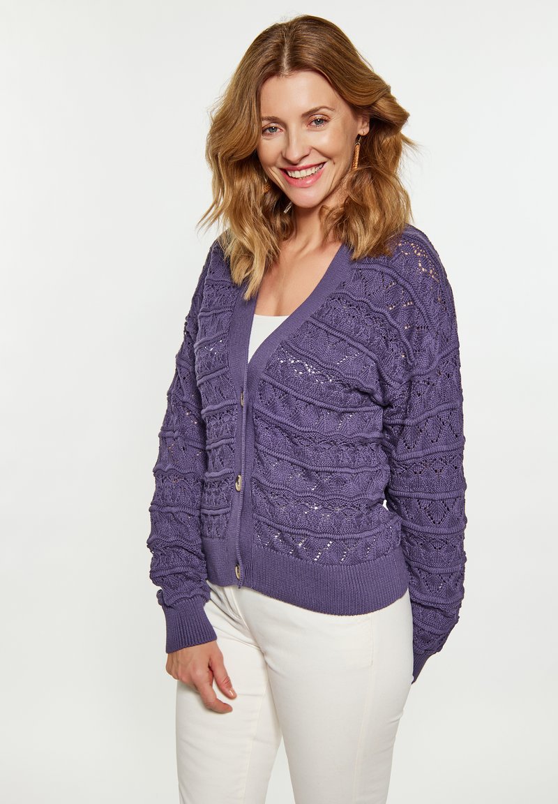 Cardigan lavorato a maglia viola con un motivo a trama, scollo a V e cinque bottoni. Presenta polsini e orlo a coste. Indossato sopra un top bianco e pantaloni chiari.