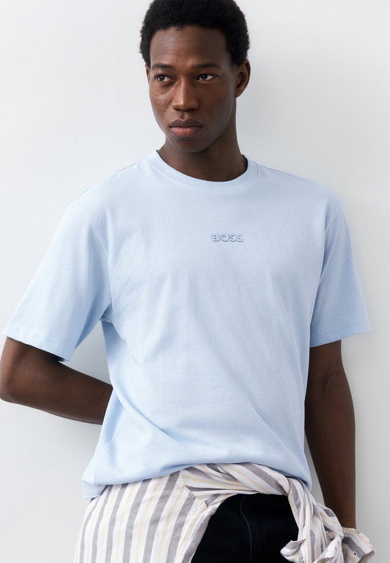 Νεαρός άνδρας με κοντά σγουρά μαλλιά, φορώντας ένα ανοιχτό μπλε T-shirt με το λογότυπο "BOSS" και ένα ριγέ πουκάμισο δεμένο γύρω από τη μέση του.