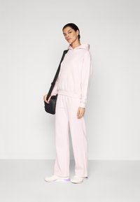 Felpa rosa con cappuccio e tasca a marsupio, abbinata a pantaloni larghi coordinati. Il modello tiene in mano una borsa a tracolla nera e indossa sneakers bianche.