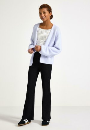 Giovane sorridente, indossa una maglietta bianca a pois, un cardigan viola chiaro, pantaloni neri e sneakers nere con strisce bianche, in piedi su uno sfondo semplice.