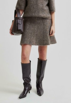 Grauer, flauschiger Strickpullover und Rock mit ausgestelltem Saum. Glänzende schwarze Lederstiefel bis zum Knie mit spitzen Zehen. Kleine, strukturierte braune Handtasche.