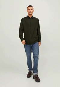 Άνδρας που περπατάει προς τα μπροστά, φορώντας ένα μαύρο πουκάμισο με κουμπιά, μπλε τζιν με διπλωμένες μπούκλες και καφέ δερμάτινα παπούτσια σε απλό φόντο.